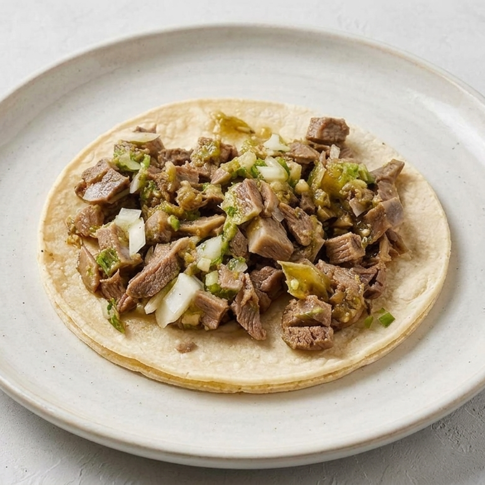 Soft corn tortillas topped with chopped grilled meat, onions, cilantro, and green salsa on a ceramic plate.